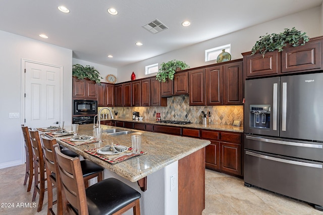 kitchen with black appliances, a kitchen bar, sink, an island with sink, and light stone counters