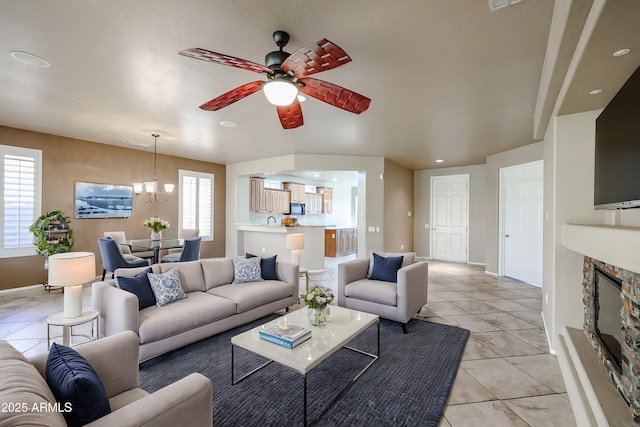 living room featuring light tile patterned floors, ceiling fan with notable chandelier, and a stone fireplace