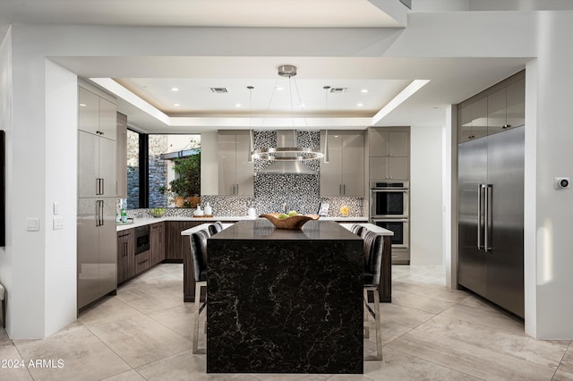 kitchen with backsplash, a spacious island, a raised ceiling, built in appliances, and wall chimney exhaust hood