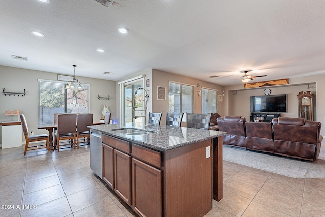 kitchen featuring pendant lighting, a center island with sink, ceiling fan with notable chandelier, stainless steel dishwasher, and dark stone countertops