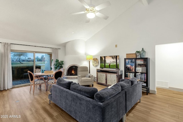 living area with a warm lit fireplace, visible vents, ceiling fan, light wood-type flooring, and high vaulted ceiling