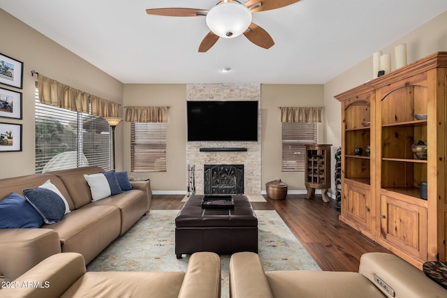 living room with ceiling fan, a stone fireplace, baseboards, and wood finished floors