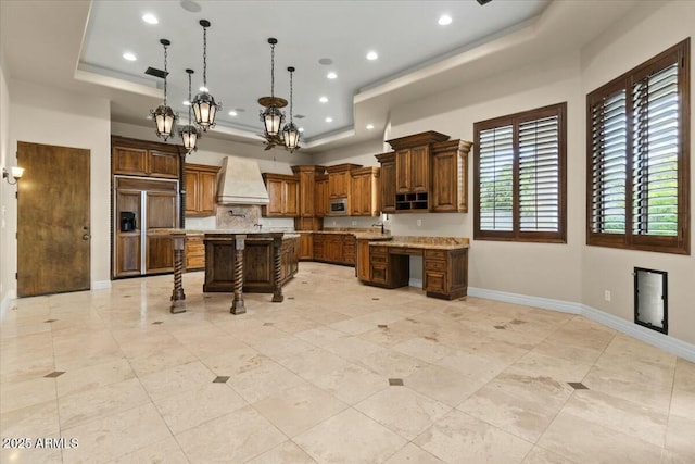 kitchen with premium range hood, a kitchen breakfast bar, a kitchen island with sink, light stone counters, and a raised ceiling