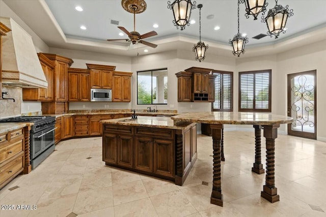 kitchen featuring pendant lighting, premium range hood, appliances with stainless steel finishes, a center island with sink, and a raised ceiling