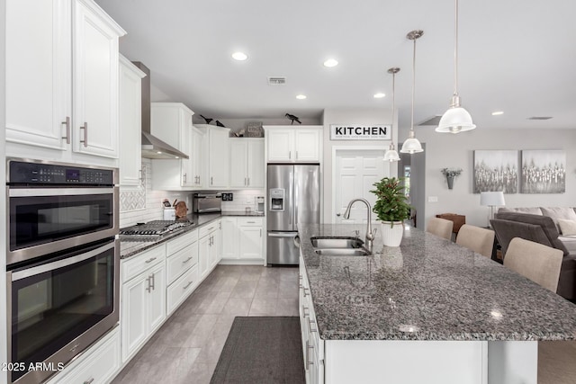 kitchen featuring wall chimney exhaust hood, stainless steel appliances, a kitchen island with sink, sink, and pendant lighting