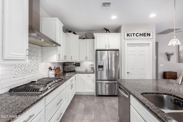 kitchen with dark stone counters, wall chimney range hood, sink, appliances with stainless steel finishes, and decorative light fixtures