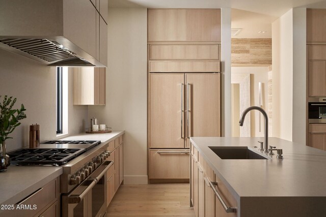 kitchen with wall chimney exhaust hood, premium appliances, light brown cabinetry, a center island with sink, and sink