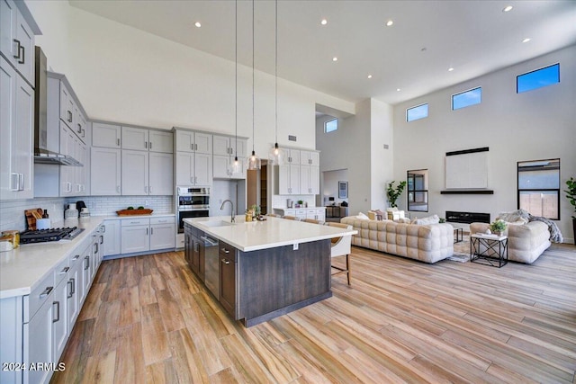 kitchen featuring appliances with stainless steel finishes, light hardwood / wood-style floors, an island with sink, decorative light fixtures, and wall chimney exhaust hood