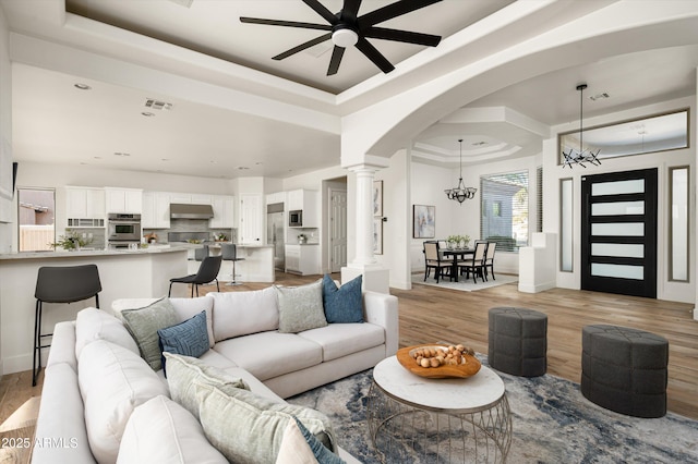 3946 N Pinnacle Hills Cir, Mesa, AZ 85207 - living room featuring decorative columns, a raised ceiling, a ceiling fan, light wood-style flooring, and a chandelier