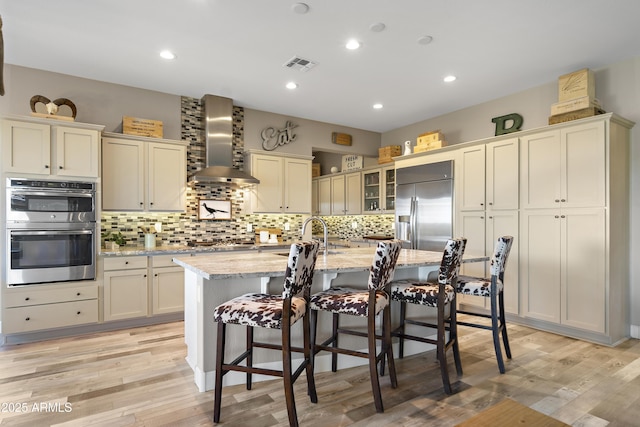 kitchen with appliances with stainless steel finishes, light stone counters, an island with sink, a kitchen bar, and wall chimney exhaust hood