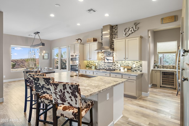 kitchen featuring sink, hanging light fixtures, light stone countertops, a kitchen island with sink, and wall chimney range hood