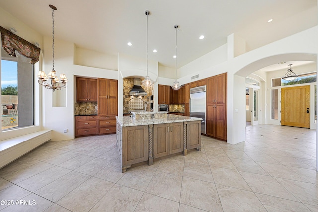 kitchen with stainless steel built in fridge, a notable chandelier, arched walkways, and wall chimney exhaust hood