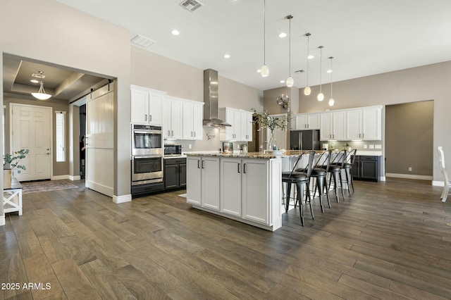 kitchen featuring a barn door, dark wood-type flooring, stainless steel double oven, wall chimney range hood, and refrigerator with ice dispenser