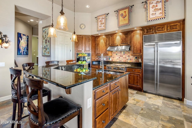 kitchen featuring decorative backsplash, a kitchen bar, dark stone counters, stainless steel built in refrigerator, and an island with sink