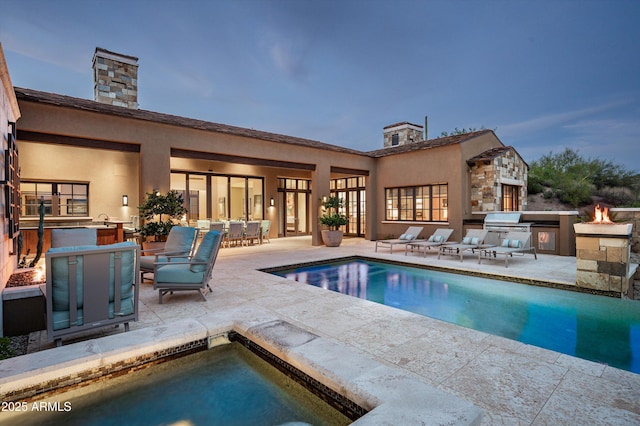 rear view of house with a chimney, exterior kitchen, an outdoor pool, a patio area, and stucco siding