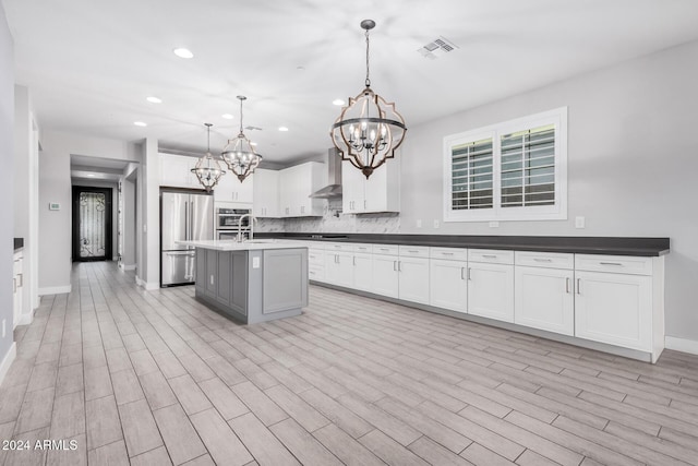 kitchen with white cabinetry, wall chimney range hood, pendant lighting, a kitchen island with sink, and appliances with stainless steel finishes