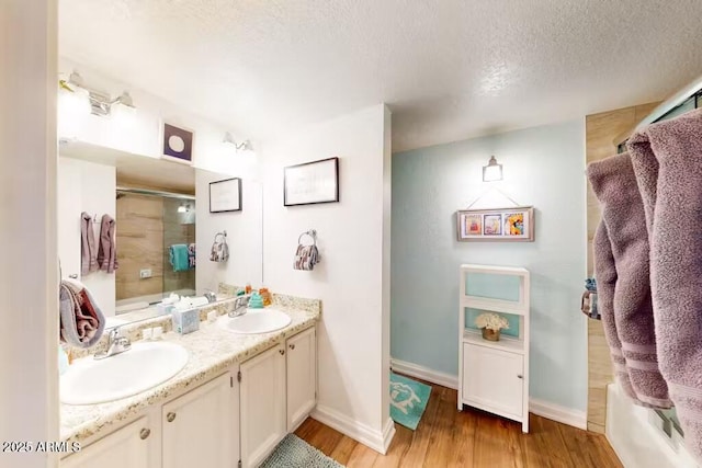 full bath featuring wood finished floors, double vanity, a textured ceiling, baseboards, and combined bath / shower with glass door