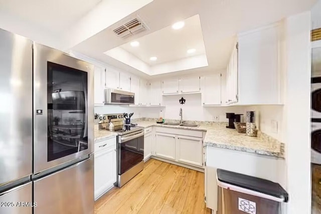 kitchen featuring appliances with stainless steel finishes, a sink, a tray ceiling, light wood finished floors, and white cabinets