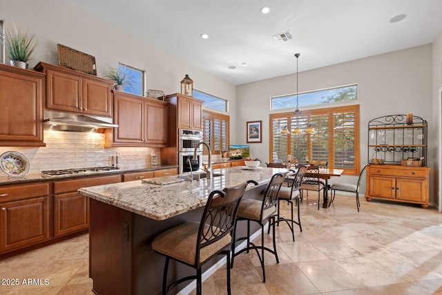 kitchen with light stone countertops, appliances with stainless steel finishes, decorative light fixtures, a breakfast bar area, and an island with sink