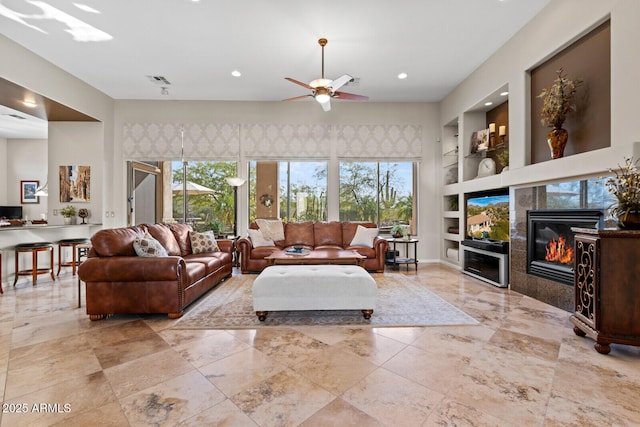 living room with a tiled fireplace, ceiling fan, built in features, and a healthy amount of sunlight