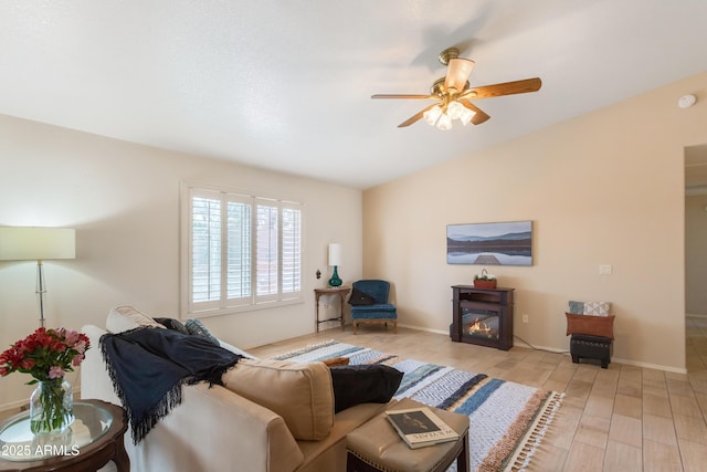 living room featuring a ceiling fan, baseboards, vaulted ceiling, light wood-type flooring, and a glass covered fireplace