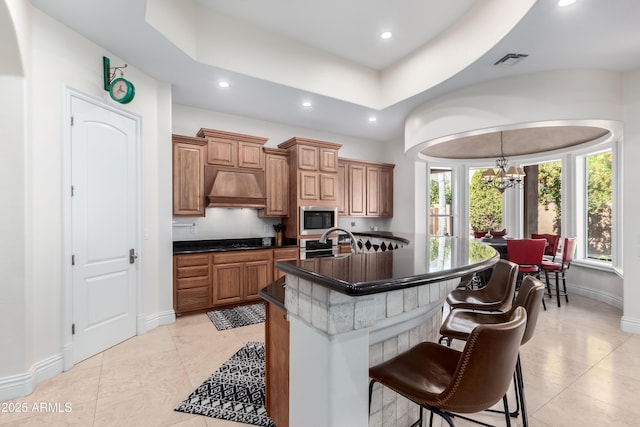 kitchen featuring stainless steel microwave, a kitchen breakfast bar, a kitchen island with sink, custom range hood, and an inviting chandelier