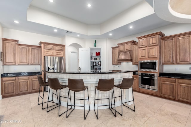 kitchen with a raised ceiling, a breakfast bar area, custom exhaust hood, a kitchen island with sink, and stainless steel appliances