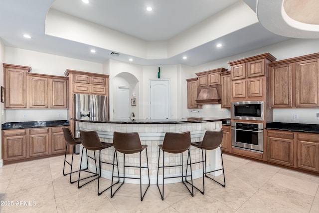 kitchen featuring premium range hood, a kitchen island with sink, stainless steel appliances, a tray ceiling, and a kitchen bar