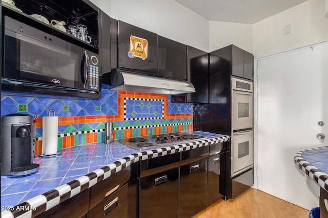 kitchen with under cabinet range hood, dark cabinets, backsplash, tile counters, and stainless steel appliances