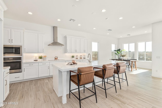 kitchen with built in microwave, a kitchen breakfast bar, stainless steel oven, a center island with sink, and wall chimney exhaust hood