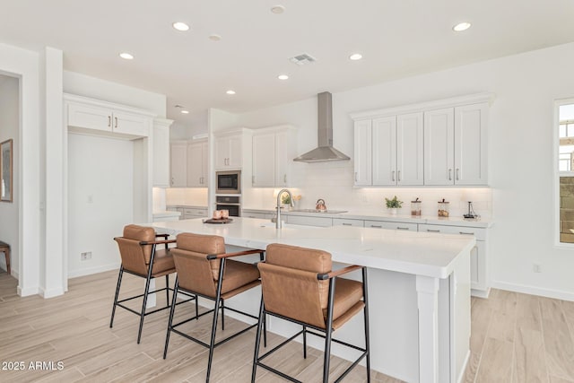 kitchen with a breakfast bar, white cabinetry, an island with sink, stainless steel oven, and wall chimney exhaust hood