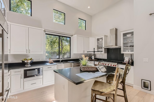 kitchen featuring a towering ceiling, wall chimney range hood, built in microwave, a sink, and light wood finished floors