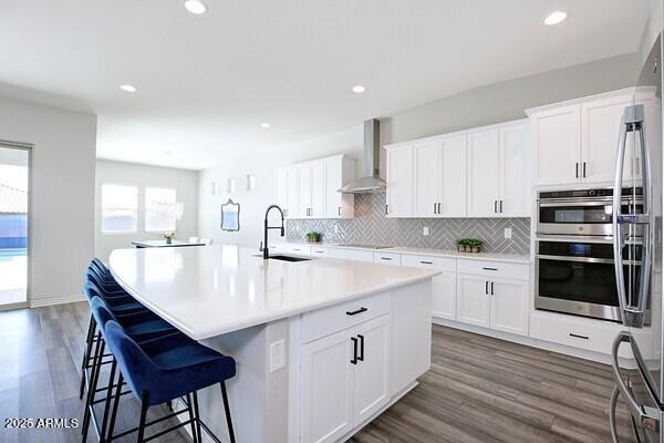 kitchen with a kitchen island with sink, a sink, dark wood-style floors, wall chimney range hood, and decorative backsplash