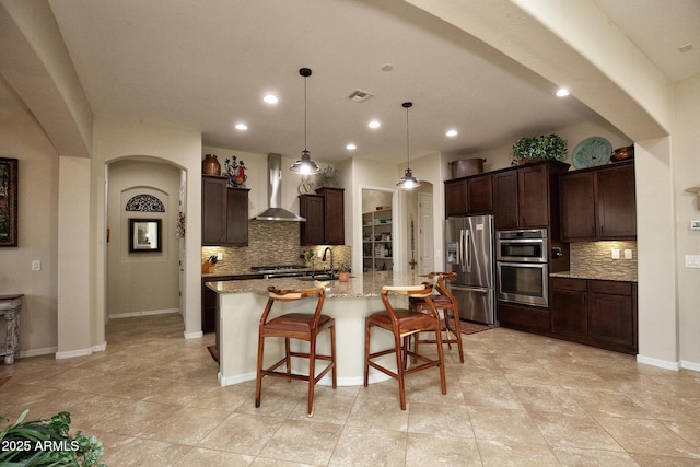 kitchen with a kitchen bar, light stone counters, stainless steel appliances, a kitchen island with sink, and wall chimney range hood