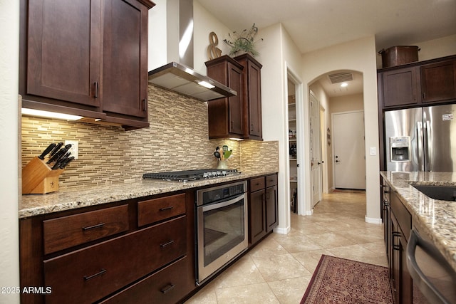 kitchen featuring light stone countertops, wall chimney exhaust hood, backsplash, light tile patterned flooring, and appliances with stainless steel finishes