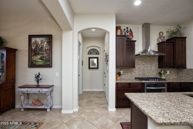 kitchen featuring backsplash, wall chimney range hood, light stone counters, dark brown cabinetry, and stainless steel appliances