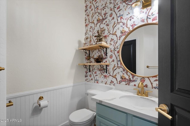 half bathroom with a wainscoted wall, toilet, vanity, and wallpapered walls