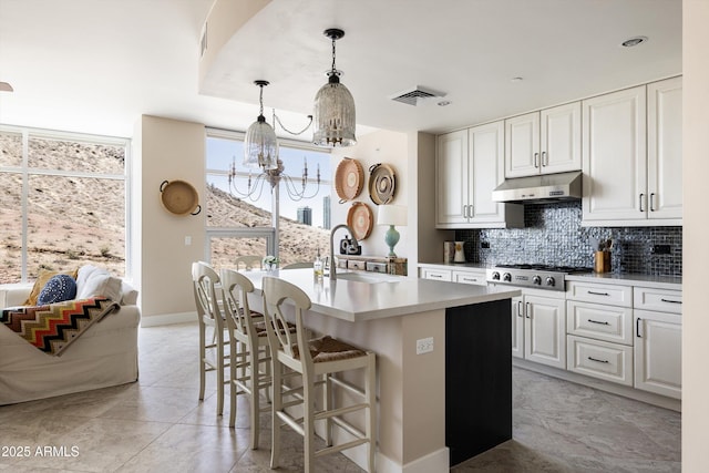 kitchen with a sink, under cabinet range hood, decorative backsplash, stainless steel gas cooktop, and a chandelier