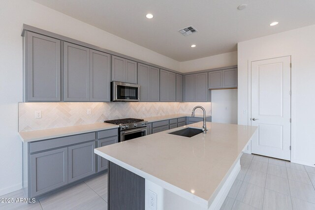 kitchen featuring sink, gray cabinets, decorative backsplash, a center island with sink, and appliances with stainless steel finishes