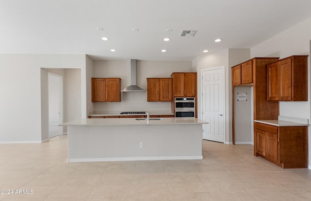 kitchen with wall chimney exhaust hood, double oven, a center island with sink, and light tile patterned floors