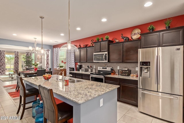 kitchen with dark brown cabinetry, light stone countertops, a kitchen island with sink, stainless steel appliances, and a sink