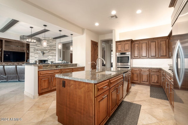 kitchen featuring a fireplace, stainless steel appliances, decorative light fixtures, dark stone countertops, and a center island with sink