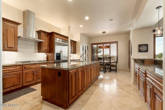 kitchen featuring a kitchen island with sink, wall chimney exhaust hood, dark stone counters, decorative light fixtures, and appliances with stainless steel finishes