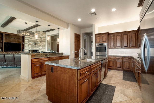 kitchen with sink, an island with sink, stainless steel appliances, dark stone counters, and decorative light fixtures