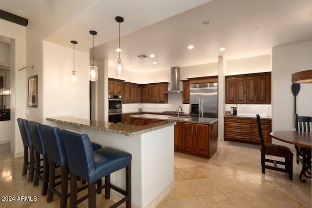 kitchen with wall chimney range hood, kitchen peninsula, dark stone counters, pendant lighting, and appliances with stainless steel finishes