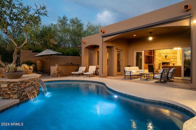 pool at dusk with pool water feature, a patio, and an outdoor stone fireplace