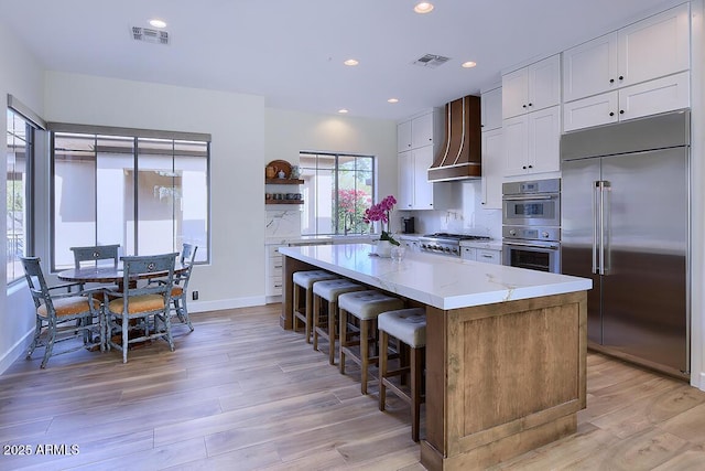 kitchen featuring a center island, wall chimney range hood, stainless steel appliances, light stone countertops, and white cabinets