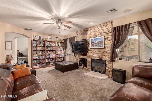 living room featuring visible vents, carpet floors, arched walkways, ceiling fan, and a stone fireplace