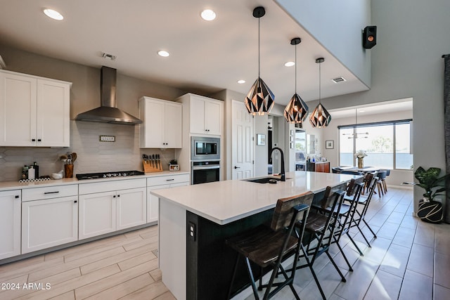 kitchen featuring wall chimney range hood, an island with sink, sink, white cabinetry, and appliances with stainless steel finishes