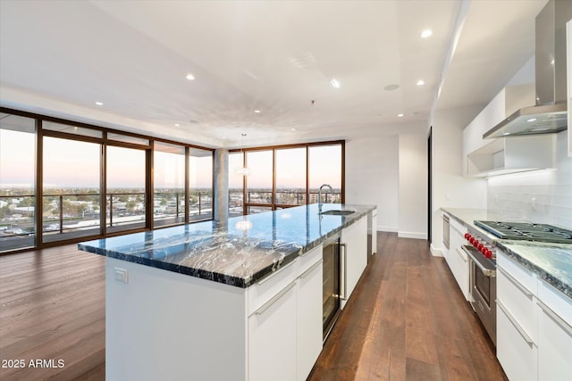 kitchen featuring dark wood-style flooring, floor to ceiling windows, a center island with sink, a sink, and wall chimney exhaust hood